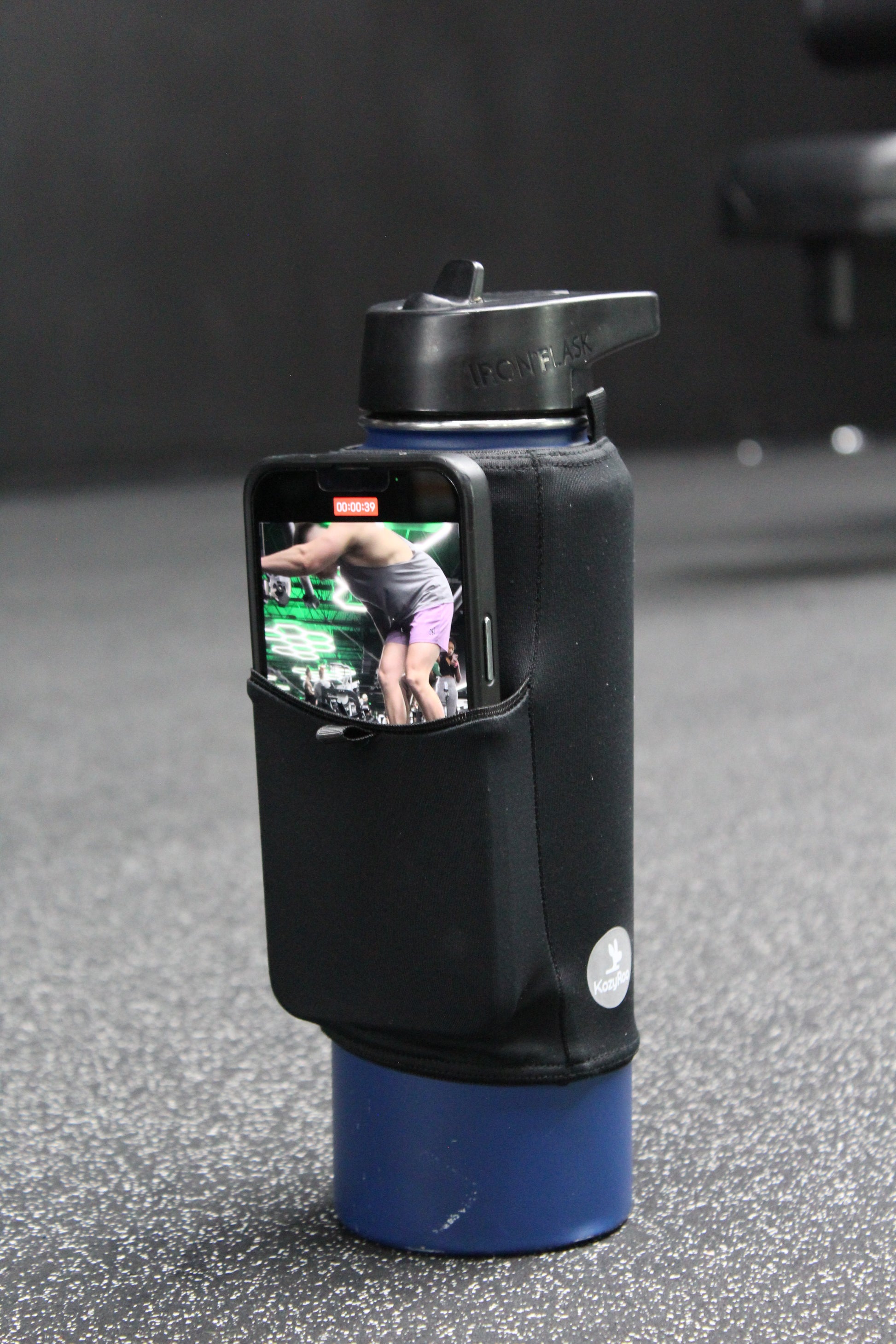 A black Kozy Sleeve on a blue water bottle recording a video of a trainer lifting at the gym.